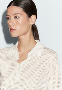 Witte, gestructureerde shirt met een gebreid patroon, met een kraag en een knoopsluiting. De stof lijkt lichtgewicht en ademend te zijn.