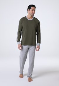 Camicia a maniche lunghe verde oliva con dettagli e polsini grigi, abbinata a pantaloni larghi morbidi e elasticizzati di un grigio chiaro. Abbigliamento casual adatto per stare a casa.