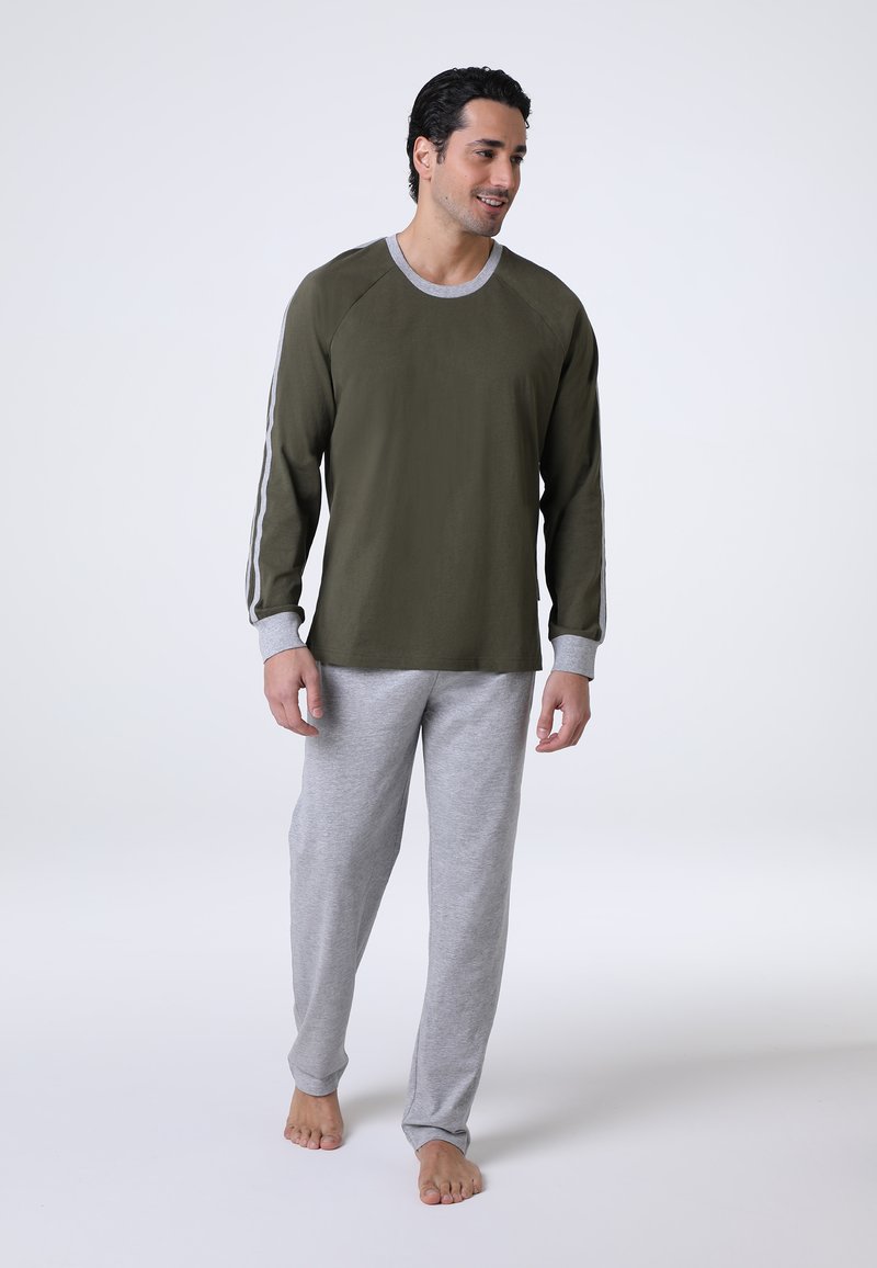 Camicia a maniche lunghe verde oliva con dettagli e polsini grigi, abbinata a pantaloni larghi morbidi e elasticizzati di un grigio chiaro. Abbigliamento casual adatto per stare a casa.