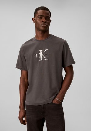 Muž v tmavosivom tričku Calvin Klein s veľkým bielym logom CK, tmavé nohavice a strieborný náramok, stojaci pred jednoduchým svetlým pozadím.
