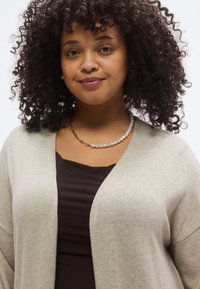 Ανοιχτό γκρι ζακέτα από απαλό, υφασμένο ύφασμα, ανοιχτό μπροστινό μέρος, συνδυασμένο με σκούρο καφέ τοπ. Εντυπωσιακό κολιέ με πέρλες και χρυσή αλυσίδα.