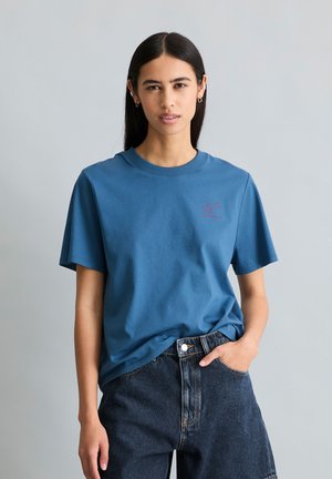 Femme aux longs cheveux foncés portant un t-shirt bleu et un jean en denim foncé, debout avec une main dans la poche devant un fond gris clair.