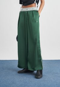 Άτομο που φοράει στενές σκούρες πράσινες φόρμες Nike με λευκό λογότυπο και ριγέ λευκή μέση, στέκεται σε μπλε χαλί με τα χέρια στις τσέπες.