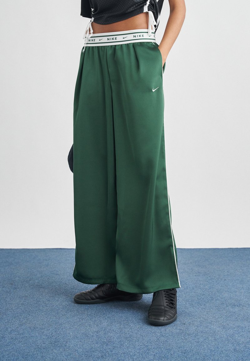 Άτομο που φοράει στενές σκούρες πράσινες φόρμες Nike με λευκό λογότυπο και ριγέ λευκή μέση, στέκεται σε μπλε χαλί με τα χέρια στις τσέπες.