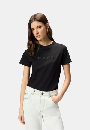 Donna con capelli castani indossa una maglietta nera a maniche corte con la scritta "PINKO" e jeans azzurri, posando con una mano in tasca.