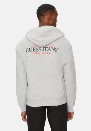 Hombre vestido con sudadera con capucha de color gris claro con el texto "GUESS JEANS" y letras rojas en la espalda, combinado con pantalones negros, de espaldas frente a un fondo blanco.
