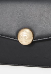 Svart texturerad läderplånbok med en gyllene rund lås som har Lacoste-logotypen, accentuerad med fina sömmar längs kanterna.