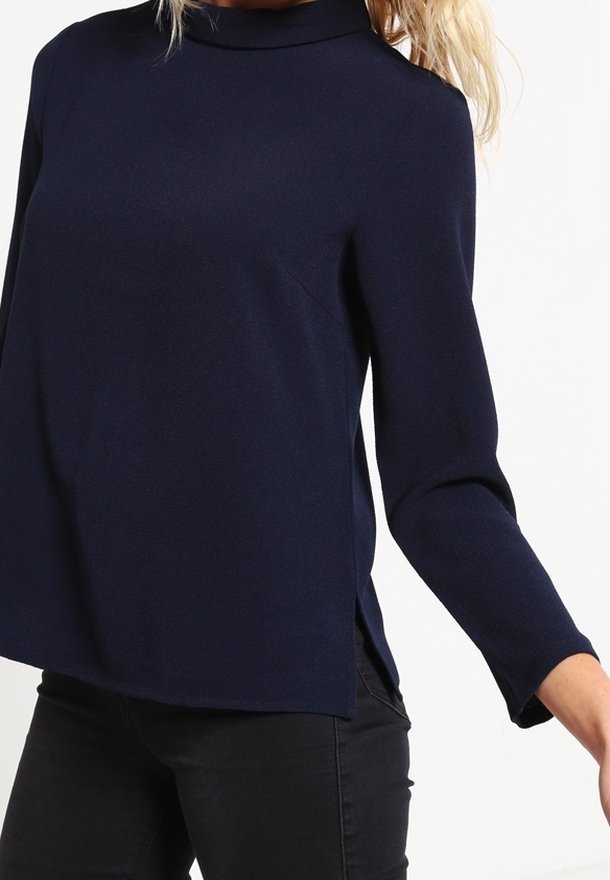 Femme portant un haut bleu marine foncé à manches longues avec un col haut et des fentes latérales, associé à un pantalon noir sur un fond uni.