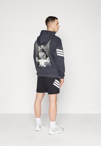 Marineblå hettegenser med grafisk design på baksiden, tre hvite striper på hver erme, kombinert med svarte shorts og lyse joggesko.