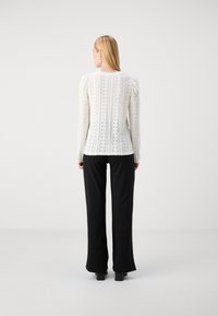 White long-sleeve top with vertical knit pattern and cut-out details, paired with black flared pants. The setting is minimalist and neutral.