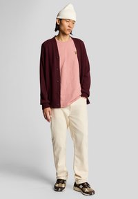 Vinrød cardigan med knapper over en lyserød kortærmet skjorte. Hvid ribstrikket hue, cremefarvede bukser og brune sneakers med mønster.