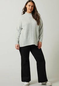 Lichtgrijze gebreide trui met een hoge nek en een getextureerd diagonaal patroon, gecombineerd met zwarte wijde broek en witte sneakers.