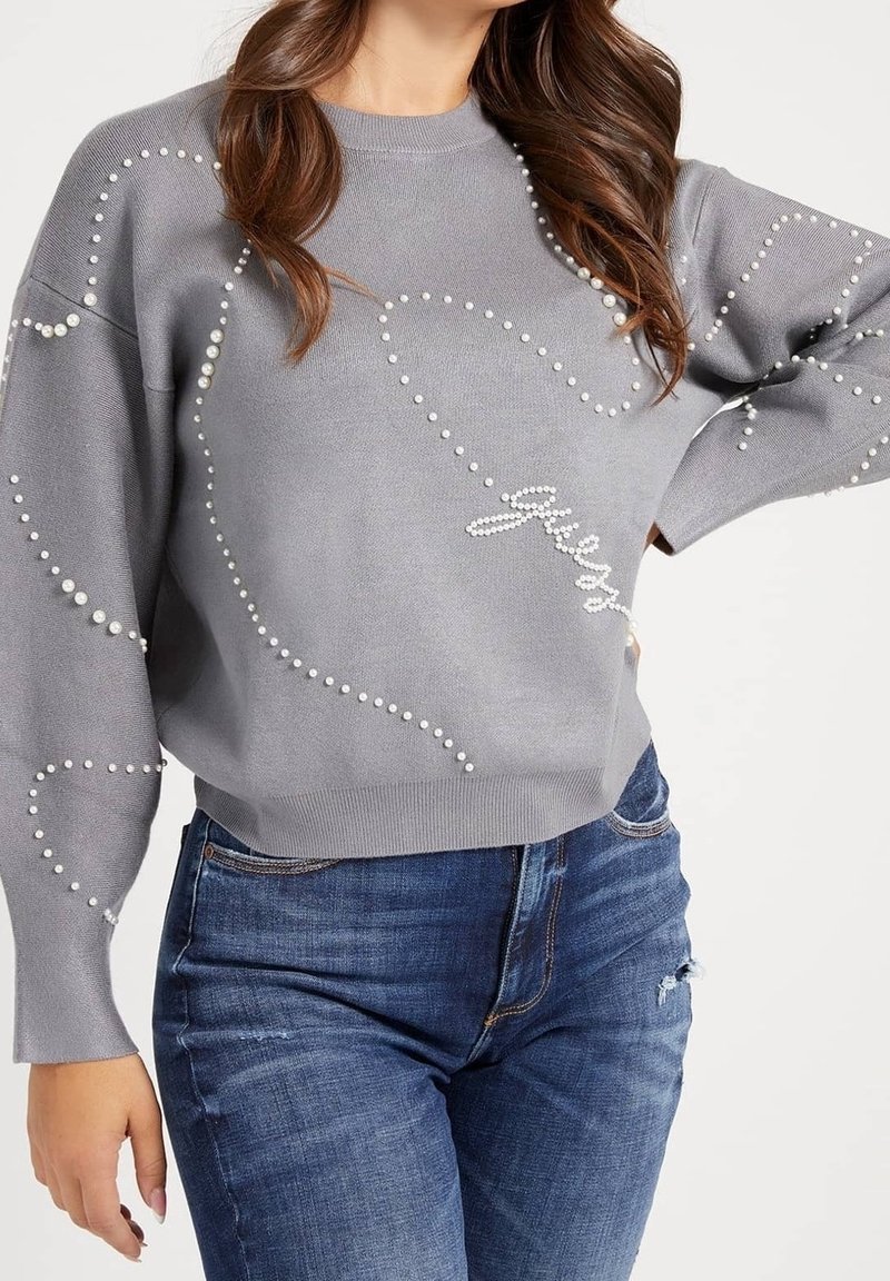 Grauer Pullover aus weichem Stoff, mit lockerem Schnitt, Perlendetails und dekorativer "Guess"-Signatur. Kombiniert mit blauen Jeans.