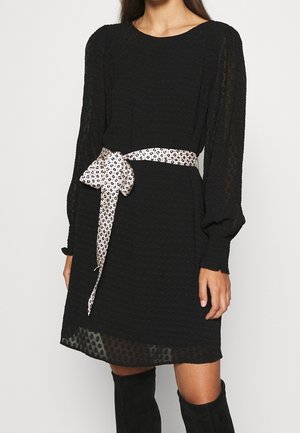 Robe noire texturée à manches longues avec manches et ourlet transparents, nouée à la taille avec une ceinture blanche et noire à motifs, portée avec des bottes noires montant aux genoux.