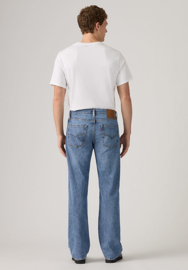 Homme portant un t-shirt blanc et un jean bleu droit, debout, dos tourné, devant un fond uni.