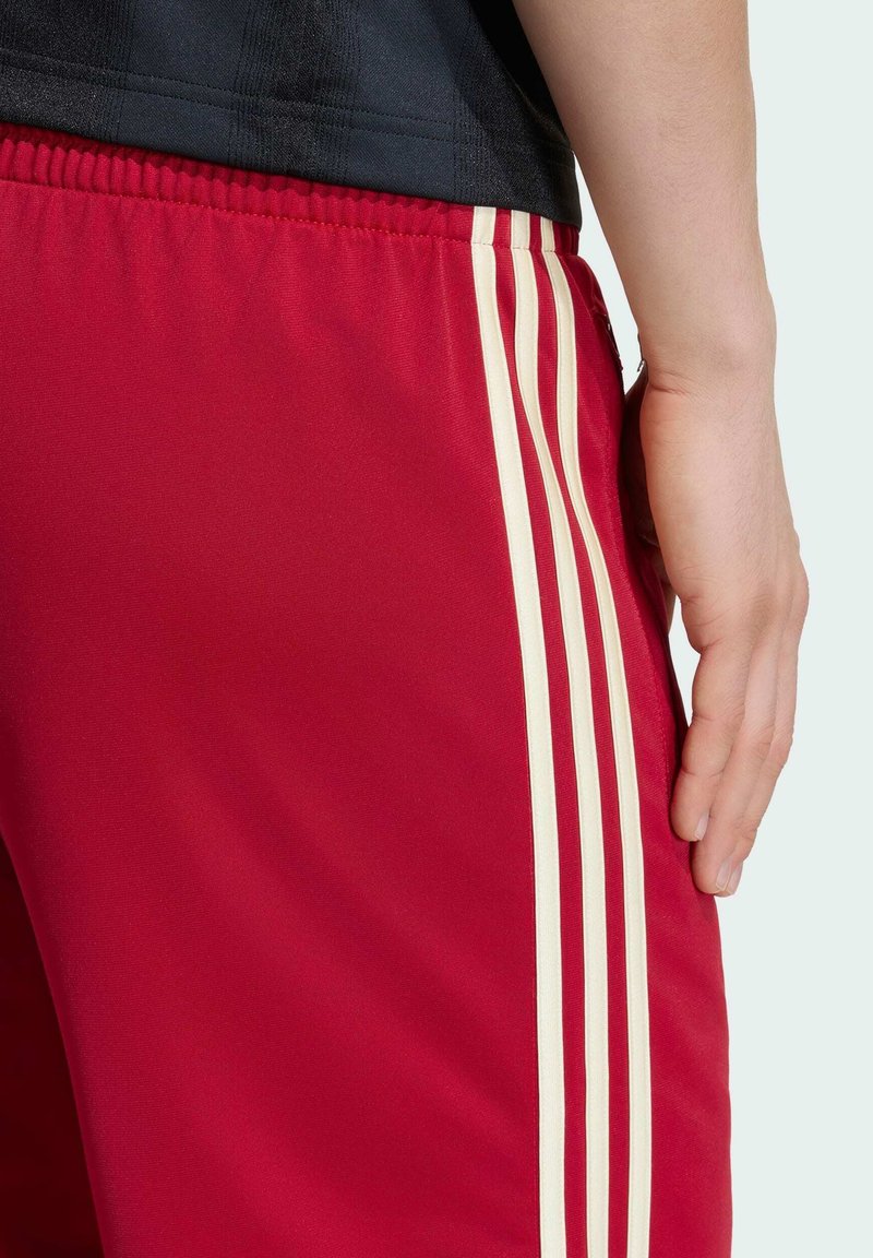Close-up af person iført røde atletikshorts med tre hvide lodrette striber og en sort skjorte, hånd afslappet ved siden.