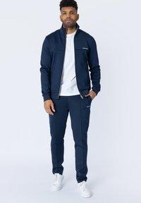Costum de sport albastru închis, cu jachetă cu fermoar și pantaloni, având buzunare laterale, logo alb accent și adidași albi. Material neted și ajustat.