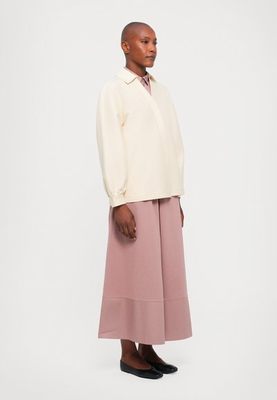 Κρεμ μπλούζα με γιακά και μακρύ μανίκι, συνδυασμένη με ροζ μακριά φούστα. Το μοντέλο φοράει μαύρες �μπαλαρίνες και στέκεται στο πλάι.