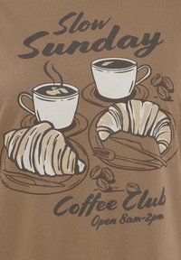 Brun t-shirt med en grafisk design av två kaffekoppar, croissanter och kaffebönor med texten: "Slow Sunday Coffee Club Öppet 8-14".