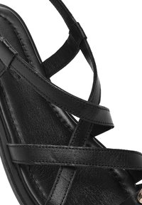 Sandalo in pelle nera con cinturini incrociati, superficie texturizzata e una leggera curvatura nella suola, con minimi dettagli decorativi.
