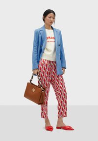Blazer blu, felpa bianca con testo rosso, pantaloni a motivi rosa e rossi, scarpe basse rosse e borsa marrone con logo "M".