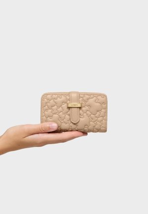 Mano che tiene un portafoglio beige con motivo floreale trapuntato e chiusura dorata, fotografato su uno sfondo grigio chiaro uniforme.