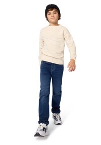 Maglione a maglia dalla texture cremosa, jeans blu e scarpe da ginnastica bianche con accenti neri. Una cuffia nera è indossata sulla testa.