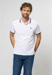 Hvid polo-shirt med rød og blå stribet krave, der har et lille sort logo på venstre bryst. Båret med blå jeans.