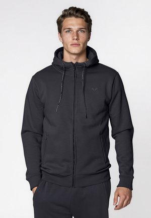 Jeune homme à la peau claire et aux cheveux bruns portant un sweat à capuche noir zippé avec cordons, debout devant un fond blanc uni.