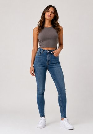Vrouw staat, draagt een grijs mouwloos crop top, hooggetailleerde blauwe jeans en witte sneakers tegen een effen achtergrond.