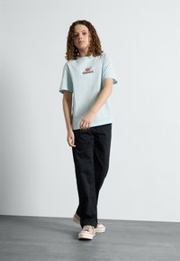 Lyseblå bomulds t-shirt med korte ærmer og et sort logo. Kombineret med løse sorte bukser og beige sneakers med røde accenter.