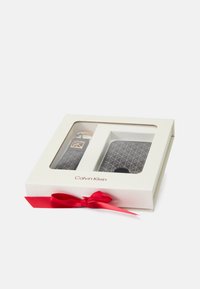Boîte cadeau Calvin Klein en blanc avec une fenêtre transparente. Contient un portefeuille à motifs et un porte-clés dans des tons sombres. Présentée avec un ruban rouge.