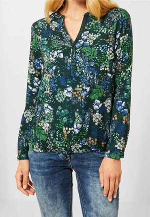 Frau trägt eine dunkelgrüne, langärmelige Bluse mit blauen, weißen und beigen Blumen, kombiniert mit blauen Jeans.