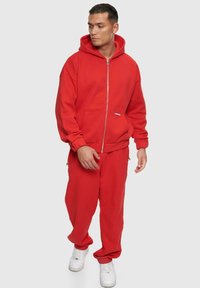 Haut à capuche zippé rouge et pantalons assortis en tissu doux. Design à capuche avec poches avant et poignets côtelés; associé à des baskets blanches.