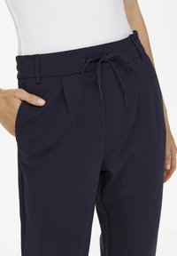 Pantalones azul marino con cintura de cordón, frente plisado, bolsillos laterales y cinturilla elástica. Hechos de tela suave con textura ligera.