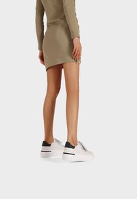 Sneakers bianchi con accenti neri, design con lacci e suola bianca spessa. Indossati con un vestito khaki, mostrando una superficie dal texture liscia.