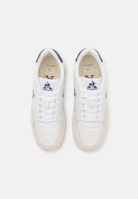 Zapatillas blancas con parte superior de cuero, detalles en beige texturizado y logo en azul marino. Incluyen cierre con cordones y puntera perforada para mayor transpirabilidad.