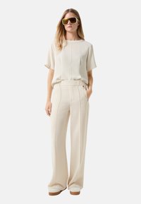 Crème-kleurig, kortmouwig shirt gecombineerd met wijde, hoge taille broek. Beide items hebben een gladde textuur en subtiele naden. Overgedimensioneerde gele zonnebril.