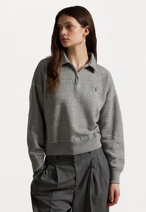 Sudadera gris con cremallera, fabricada en una tela suave con dobladillo acanalado, que presenta un pequeño logo azul en el pecho y un corte relajado.