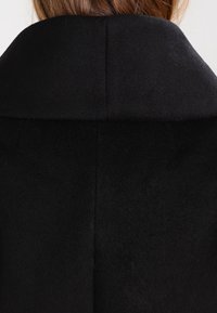 Schwarzer Mantel mit hohem Kragen, weicher Textur und nahtlosem Design. Der Stoff wirkt plüschig und steigert die Wärme und den Komfort.