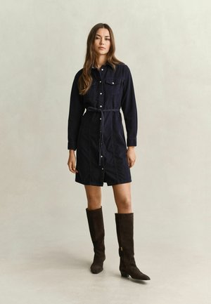 Frau stehend, trägt ein dunkles, langärmeliges, geknöpftes Kleid mit Taillengürtel und kniehohe dunkelbraune Stiefel vor schlichtem Hintergrund.