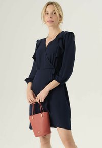 Námorníckomodr dress s volánovými rukávmi, krížovým strihom a priliehavým pásom. V kombinácii s malou textúrovanou červenou kabelkou so zlatými akcentmi.