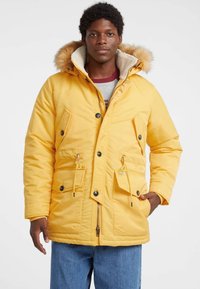 Geel geïsoleerde parka met een bontgevoerd capuchon, rits aan de voorkant, knoopsluitingen en meerdere zakken; gladde textuur en warm, gevoerd ontwerp.
