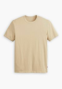 Camiseta de manga corta beige hecha de algodón suave. Cuello redondo, corte recto y una pequeña etiqueta de logo en la parte inferior izquierda. Textura suave.