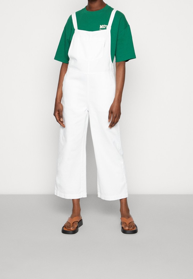 Personne portant une salopette blanche à jambes larges, un t-shirt vert à manches courtes et des sandales marron à enfiler, debout sur un sol gris.