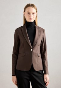 Hnědý střihový blazer s jedním knoflíkem, klopovými límci a dvěma předními kapsami. Nosený přes černý rolák. Hladká tkanina.