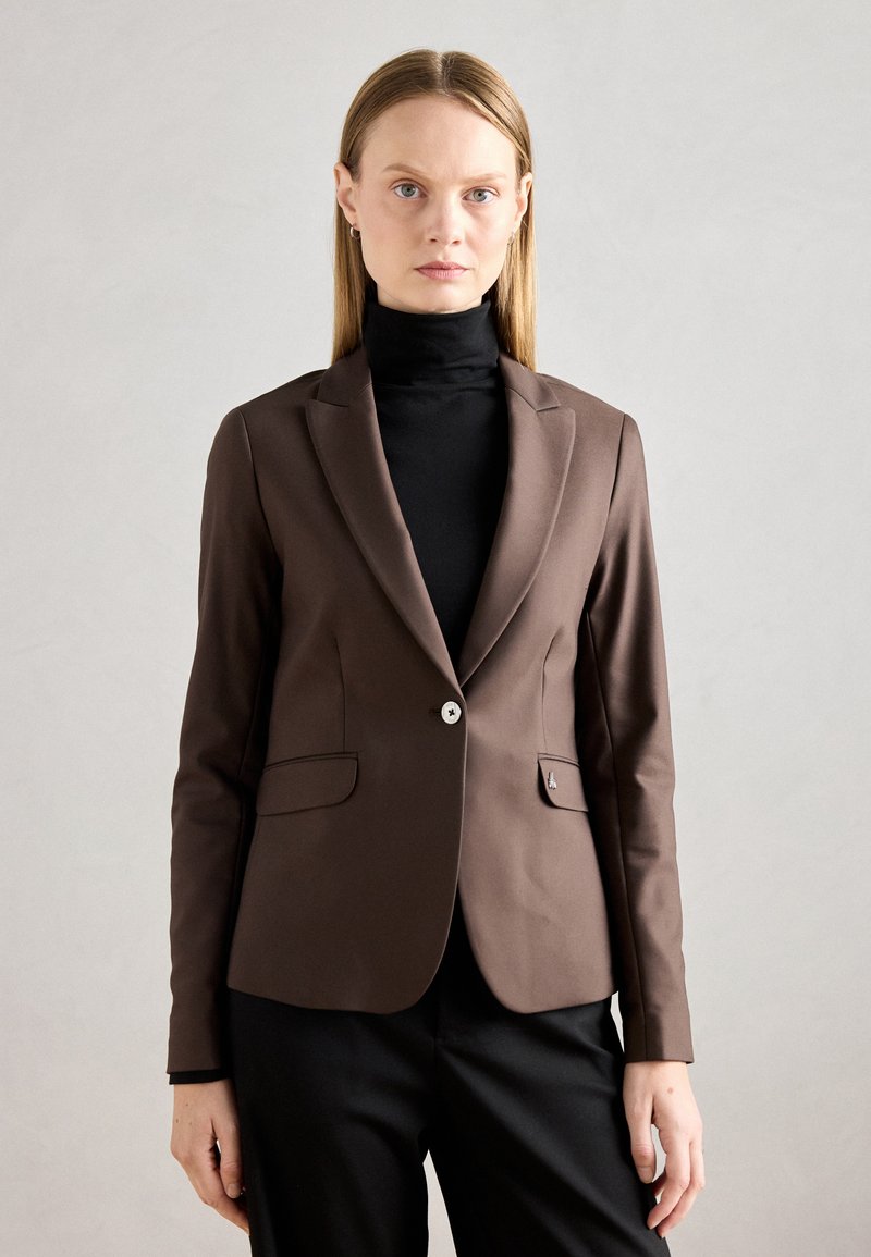 Hnědý střihový blazer s jedním knoflíkem, klopovými límci a dvěma předními kapsami. Nosený přes černý rolák. Hladká tkanina.