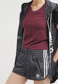 Burgundy kortärmad skjorta tillsammans med svarta mönstrade shorts med vita ränder, ficka med dragkedja och en Adidas-logotyp på fållen.