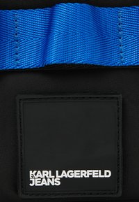 Borsa nera con superficie testurizzata, tracolla blu intrecciata e un'etichetta rettangolare nera con testo bianco "KARL LAGERFELD JEANS."