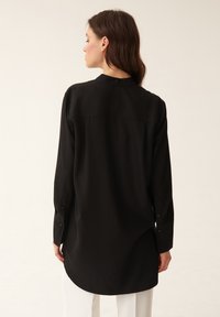 Schwarzes Langarmshirt aus glattem Stoff, mit abgerundetem Saum und geknöpften Bündchen. Die Rückansicht zeigt ein schlichtes, tailliertes Design.